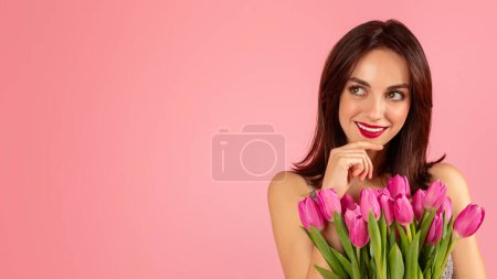 Photo for Contemplative woman with a soft smile, resting her chin on her hand, holding a bouquet of bright pink tulips, wearing a sequin dress, with a pink backdrop - Royalty Free Image