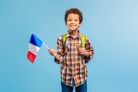 Téléchargez les photos : Joyeux garçon tenant le drapeau français, vêtu d'une chemise à carreaux avec sac à dos, incarnant l'esprit jeune et la fierté culturelle sur fond bleu - en image libre de droit