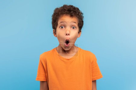 Photo for Amazed young boy with curly hair, wide-eyed in shock, wearing an orange t-shirt against calming blue background, capturing moment of astonishment - Royalty Free Image