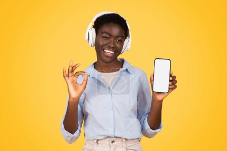 Téléchargez les photos : Heureuse jeune étudiante afro-américaine en casque sans fil, écouter de la musique, montrer smartphone avec écran vide, profiter de leçons en ligne, isolée sur fond de studio jaune. Études et éducation - en image libre de droit