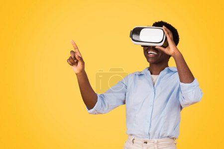 Téléchargez les photos : Glad teen black lady student in VR glasses pointe du doigt l'espace libre, l'expression de l'émerveillement et de la curiosité, isolé sur fond jaune, studio. Etude, innovation dans l'éducation - en image libre de droit