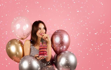 Foto de Una mujer pensativa con una sonrisa sutil, sosteniendo una pila de rosquillas encendida por velas, está rodeada de globos metálicos y confeti cayendo sobre un fondo rosa festivo. Evento de celebración - Imagen libre de derechos