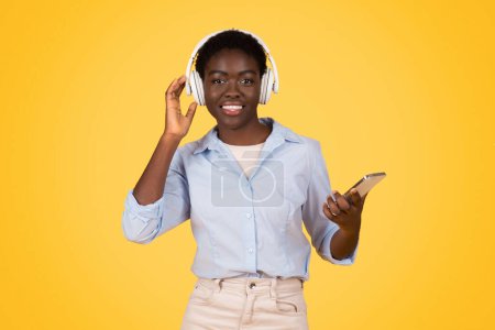 Photo for Cheerful millennial african american lady student in wireless headphones, listen music, use phone, enjoy online lesson, isolated on yellow studio background. Study and education app - Royalty Free Image