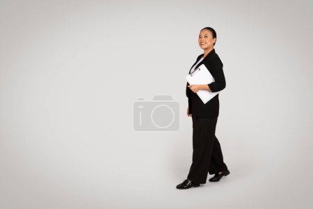 Téléchargez les photos : Souriant confiant jeune femme asiatique en costume avec presse-papiers marche, isolé sur fond gris, studio. Entreprise prospère, démarrage, travail moderne et style de vie professionnel - en image libre de droit