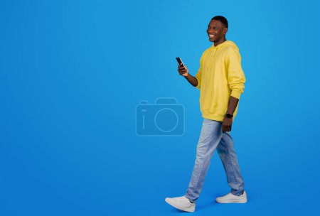 Photo for Smiling African American man student in a casual yellow hoodie and blue jeans walking and looking at his phone on a vibrant blue studio background, full length, ad, offer and sale - Royalty Free Image