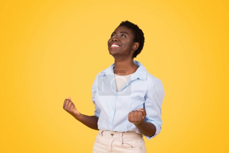 Photo for Happy teen black lady student, exuding triumph and joy, raises fists in victorious gesture, isolated on yellow background, studio. Celebrate success and win, study, work, education - Royalty Free Image
