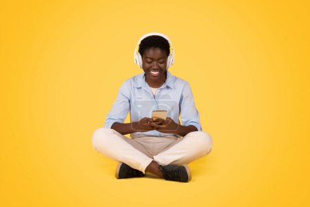 Téléchargez les photos : Jeune étudiante noire souriante dans des écouteurs bavarder au téléphone, écouter de la musique, se détendre, s'asseoir sur le sol, isolé sur fond jaune, studio. Dame profiter du repos, temps libre - en image libre de droit