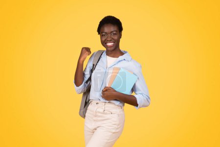 Foto de Adolescente afro-americana positiva estudiante con libros, exudando triunfo y alegría, levanta el puño en gesto victorioso, aislado sobre fondo amarillo, estudio. Celebrar el éxito y ganar, estudiar - Imagen libre de derechos