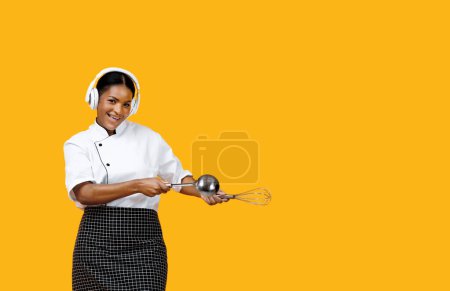 Foto de Enérgica cocinera negra bailando alegremente con cuchara y batidor en las manos, cocinera afroamericana en auriculares inalámbricos de pie aislada sobre fondo amarillo, mostrando la diversión de cocinar - Imagen libre de derechos