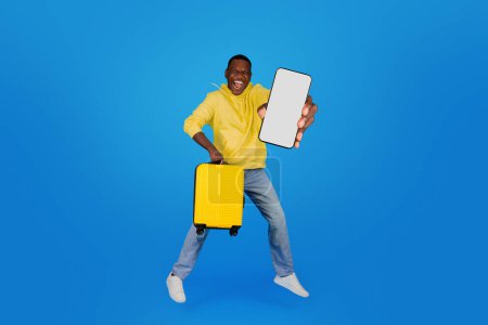 Un homme noir extatique avec une valise jaune et un smartphone avec un écran vide saute en l'air, dépeignant l'excitation du voyage tech-savvy sur un fond bleu.