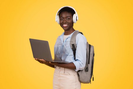 Photo for Glad young african american lady student in wireless headphones, use laptop, enjoy online lesson, isolated on yellow studio background. Study and education, listen music app - Royalty Free Image