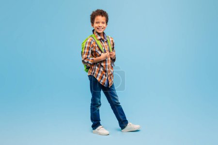 Photo for Happy schoolboy in plaid shirt and jeans with green backpack, standing confidently with smile against refreshing blue background - Royalty Free Image