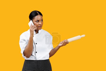 Photo for Concentrated black female chef holding rolling pin and talking on cellphone, confident african american cook woman standing against bright yellow background, depicting multitasking in busy kitchen - Royalty Free Image