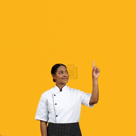 Foto de Alegre chef negra con chaqueta blanca apuntando hacia arriba en el espacio de copia por encima de su cabeza, insinuando una nueva idea o concepto, posando sobre un fondo amarillo brillante, ilustrando la creatividad - Imagen libre de derechos