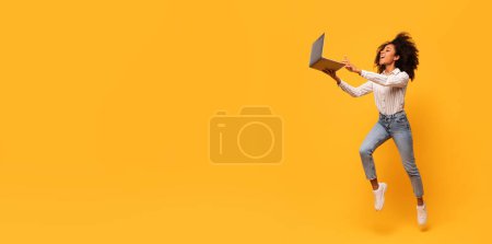 Téléchargez les photos : Jeune femme noire énergique aux cheveux bouclés bondissant joyeusement tout en tenant un ordinateur portable, symbolisant l'excitation ou le succès, sur fond jaune, panorama avec espace libre - en image libre de droit