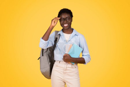 Téléchargez les photos : Une étudiante noire érudite avec des lunettes, profondément engagée dans la lecture de livres, son comportement concentré et sa curiosité intellectuelle vivement capturée sur le fond jaune vif - en image libre de droit