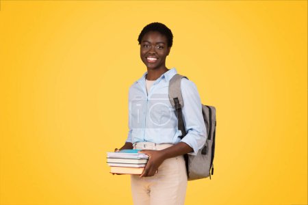Téléchargez les photos : Jeune étudiante afro-américaine positive avec livres, sac à dos, profiter du mode de vie scolaire, isolé sur fond jaune, studio. Éducation, études, publicité et offre, se préparer à l'examen - en image libre de droit