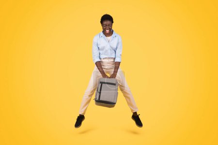 Téléchargez les photos : Joyeux jeune étudiante afro-américaine en saut décontracté avec sac à dos, à bouche ouverte, profiter du temps libre, isolé sur fond de studio jaune. Étude et éducation, amusement - en image libre de droit