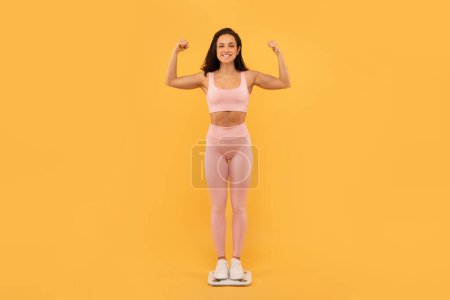 Photo for Weight Loss Success. Proud Young Fitness Lady Standing On Scales And Showing Biceps Muscles Over Yellow Studio Backdrop, Smiling To Camera, Showing Great Slimming Result. Full Length - Royalty Free Image