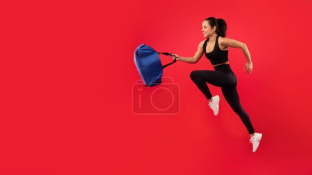 Téléchargez les photos : Jeune femme sportive en tenue de sport noire bondissant tenant sac de fitness sur fond de studio rouge, vue latérale, prise de vue pleine longueur. Sportswoman fonctionne dans la publicité en plein air gymnase promo. Panorama, espace libre - en image libre de droit