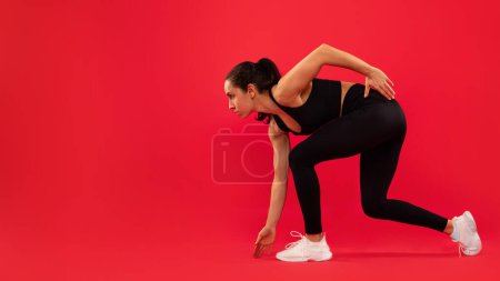 Side View Of Motivated European Lady Runner Ready For Race Standing In Crouch Start Position, Preparing For Running Over Red Studio Backdrop, Looking Aside At Copy Space. Panorama