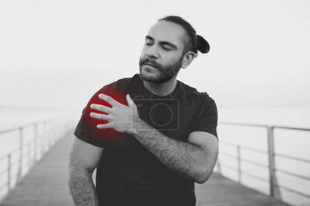 Photo for Black and white shot of young athlete man in sportswear clutches his shoulder with red pain spot during an outdoor workout at seaside, embodying the struggle of sports injuries and issues - Royalty Free Image
