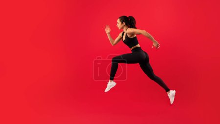 Téléchargez les photos : S'adapter jeune femme en noir vêtements de sport saute énergiquement sur fond rouge vif, incarnant la puissance et le mouvement. Vue latérale d'une sportive qui court en plein air. Panorama, espace de copie - en image libre de droit