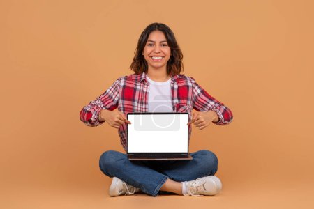 Happy middle eastern woman in casual sit on floor, point fingers at laptop with blank screen, isolated on colorful background. Blogger recommendation and device for work, study, great ad and offer