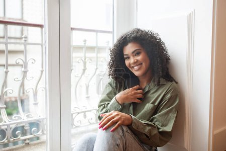 Téléchargez les photos : Jeune femme brésilienne profite d'un moment de détente assis près de la fenêtre intérieure, souriant à la caméra tout en posant dans des vêtements décontractés modernes dans ses appartements à la maison, rayonnant de bonheur - en image libre de droit