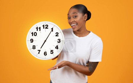 Photo for Smiling African American lady holds big clock, showcasing punctuality and deadline management, posing over orange studio backdrop, smiling to camera, embodying the concept of time - Royalty Free Image