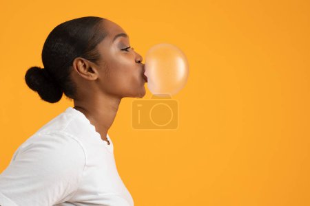 Téléchargez les photos : Profil de la dame afro-américaine souffle bubblegum pour le plaisir sur fond orange jaune vif. Restez insouciant concept. Bannière publicitaire avec espace libre pour le texte - en image libre de droit