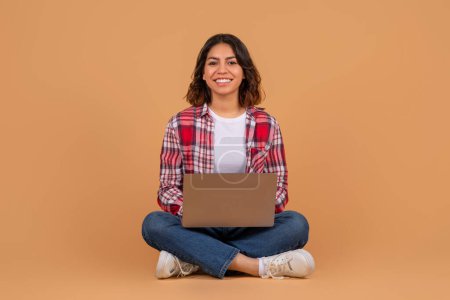 Portrait of confident young arabic woman holding and using laptop computer isolated over colorful studio background with free copy space. Smiling lady sitting on floor with pc, looking at camera