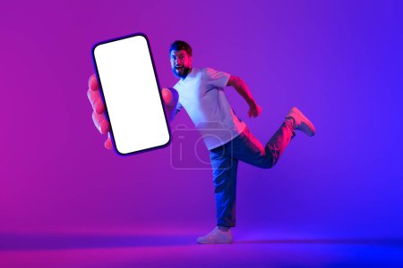 Photo for Excited Guy Shows Huge Smartphone With Blank Screen Recommending New Application, Running Over Neon Purple Blue Studio Background, Offering Cellphone Mockup For Promo Design. Collage - Royalty Free Image