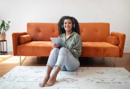 Téléchargez les photos : Jeune femme noire utilise tablette numérique portant casque sans fil, profiter de l'éducation en ligne et de divertissement, assis sur le sol dans un cadre domestique à l'intérieur. Technologie à la maison - en image libre de droit