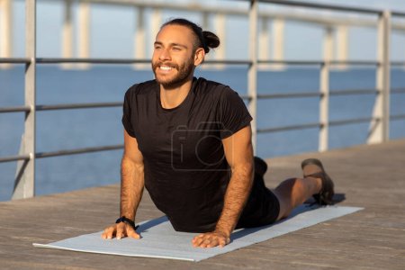 Téléchargez les photos : Homme athlétique heureux en vêtements de sport étirant les muscles du dos dans la pose de cobra, pratiquant le yoga au bord de la mer à l'extérieur, l'exercice sur tapis de fitness. Flexibilité, bien-être et forme physique - en image libre de droit