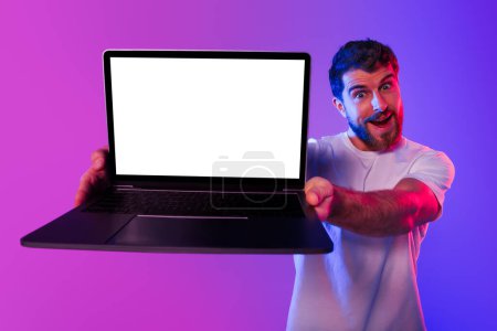 Photo for Wow online offer. Excited Guy Gamer Showing Laptop With Blank Screen Over Neon Purple Blue Studio Background, Advertising Website Template. Internet Technology Concept. Mockup - Royalty Free Image