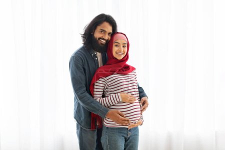 Foto de Retrato de feliz pareja musulmana embarazada posando cerca de la ventana en casa, amando al joven esposo árabe abrazando a su esposa embarazada en hijab por detrás, abrazando tiernamente su vientre, espacio para copiar - Imagen libre de derechos