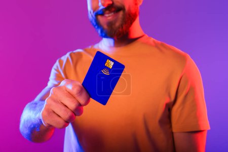 Foto de Millennial hombre mostrando azul tarjeta de crédito de plástico sobre fondo de neón púrpura, tiro recortado. Tipo publicidad banca virtual, opciones de pago ilimitadas y servicios financieros en línea - Imagen libre de derechos