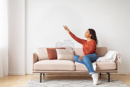 Photo for Smiling Black Woman Holding Remote Controller, Opening Air Conditioner At Home While Relaxing On Comfortable Couch In Living Room, Happy African American Female Adjusting Temperature Mode, Copy Space - Royalty Free Image