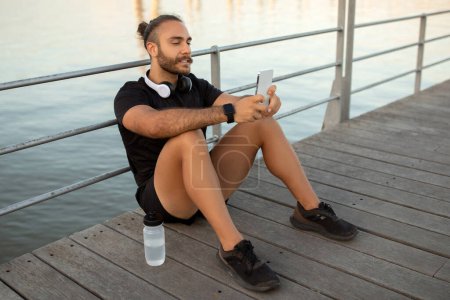 Millennial guy in sportswear enjoys seaside vibes checking fitness apps on smartphone with headphones around his neck, sitting at sea boardwalk outside, blending music, motivation and weight loss