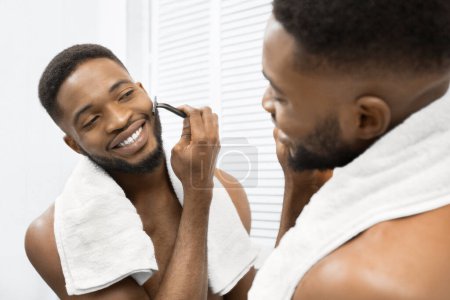 Foto de Hombre afro sonriente con el torso desnudo afeitándose la barba mirando en el espejo en el baño. Concepto de cuidado matutino - Imagen libre de derechos