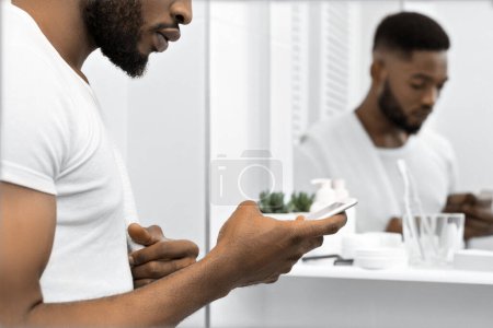 Téléchargez les photos : Beau afro homme lisant le message dans le smartphone à la salle de bain. Homme dans la salle de bain concept - en image libre de droit