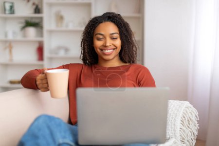 Foto de Pasatiempo en casa. Joven mujer negra descansando con café y computadora portátil en el sofá, retrato de una hermosa dama afroamericana sonriente relajándose en el sofá en la sala de estar, disfrutando del ocio de fin de semana - Imagen libre de derechos