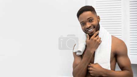 Foto de Hombre afro sonriente con el torso muscular desnudo tocando su barba en el baño. Mans concepto de cuidado diario, espacio de copia - Imagen libre de derechos