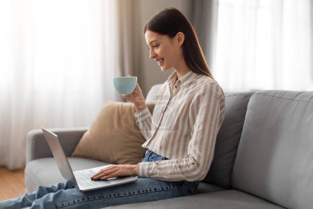 Téléchargez les photos : Jeune femme pigiste profiter d'une tasse de café tout en utilisant et en travaillant sur un ordinateur portable, assis sur un canapé gris dans un cadre lumineux et aéré salon - en image libre de droit