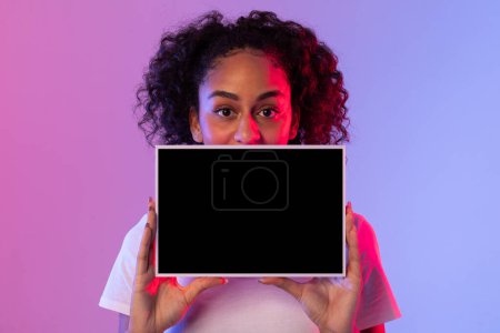 Photo for Engaging black woman with curly hair presenting horizontal tablet with black screen, perfect for mockup, against gradient neon pink and blue background - Royalty Free Image