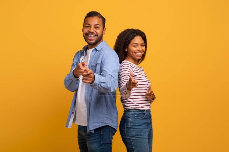 Fröhliches junges schwarzes Paar, das mit selbstbewusstem Lächeln spielerisch in die Kamera zeigt, glückliche afrikanisch-amerikanische Ehepartner aus dem Millennium, die gemeinsam in lässiger Umgebung vor leuchtend gelbem Hintergrund posieren