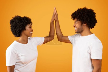 Foto de Positivo elegante rizado millennial afroamericano hombre y mujer con camisetas blancas que se dan cinco altos y sonriendo, saludándose con éxito, fondo de estudio naranja - Imagen libre de derechos