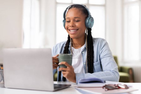 Photo for Happy black female student with braided hair wearing headphones, holding mug and studying from laptop in cozy, light-filled room, exuding relaxed vibe - Royalty Free Image