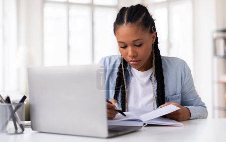 Konzentrierte schwarze Studentin mit Zöpfen, die intensiv lernt, am Schreibtisch mit Laptop in Notizbuch schreibt und im hellen Raum akademischen Fleiß verkörpert